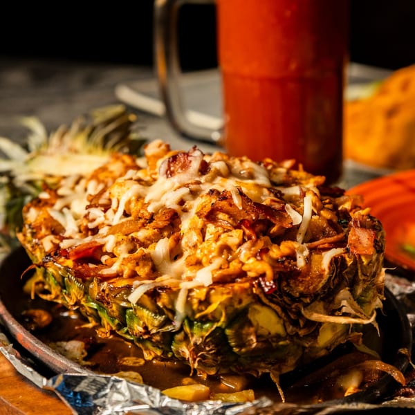 Pineapple half filled with grilled chicken and melted cheese, served with a red drink.