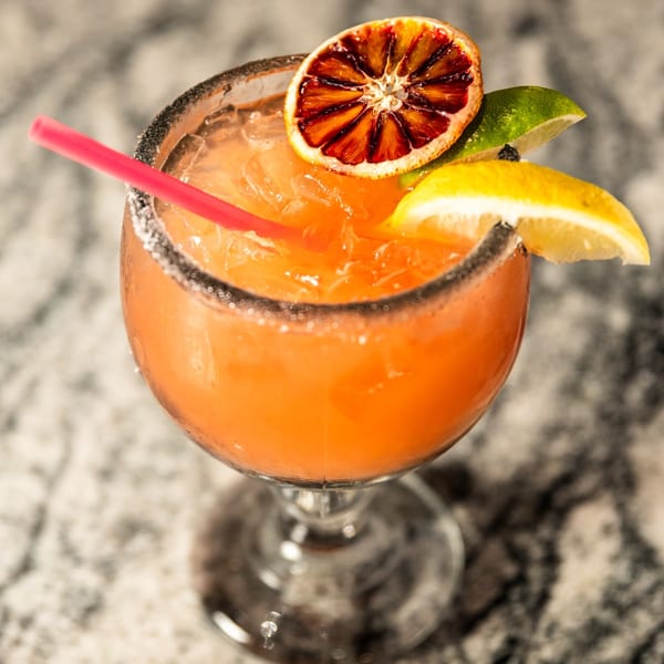 Orange cocktail with a black sugar rim, garnished with blood orange, lime, and lemon slices.