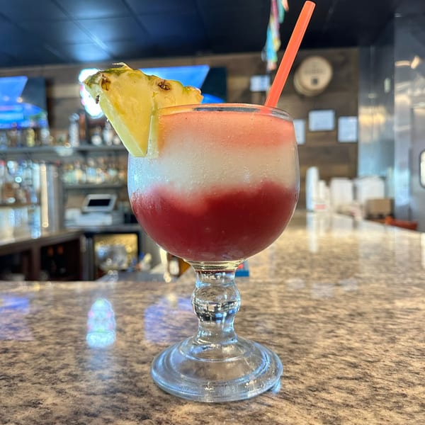 Layered red, white, and pink frozen drink with a pineapple slice and orange straw on a bar.