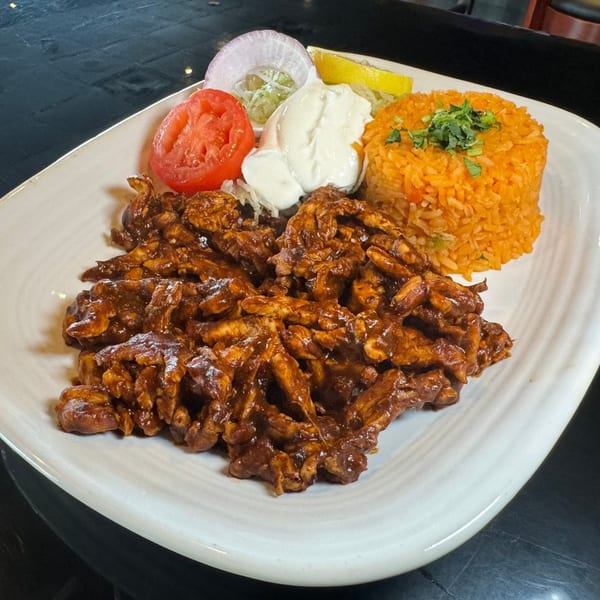 A plate of saucy chicken, orange rice, tomato, onion, sour cream, and lemon.